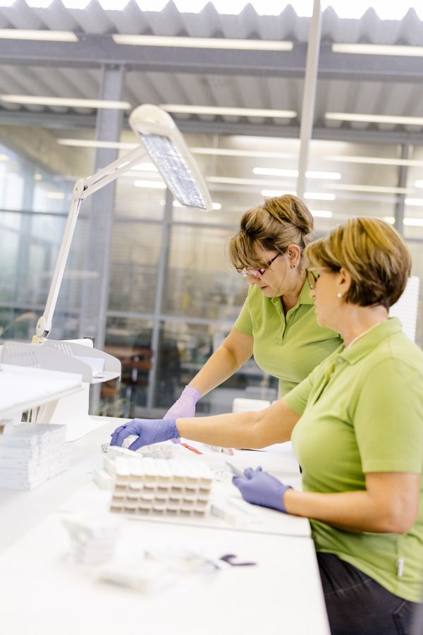 Two employees wearing green polo shirts and purple protective gloves work intently at a brightly lit table in the laboratory.