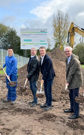 Sechs Männer stehen auf einer Baustelle und führen einen symbolischen Spatenstich durch. Im Hintergrund sind Erdhügel, ein Bagger und ein Bauschild zu sehen.