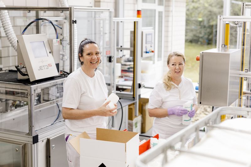 Zwei Mitarbeiterinnen der Alfred Becht GmbH stehen an der Abfüll- und Verpackungslinie und bereiten Produkte für den Versand vor.