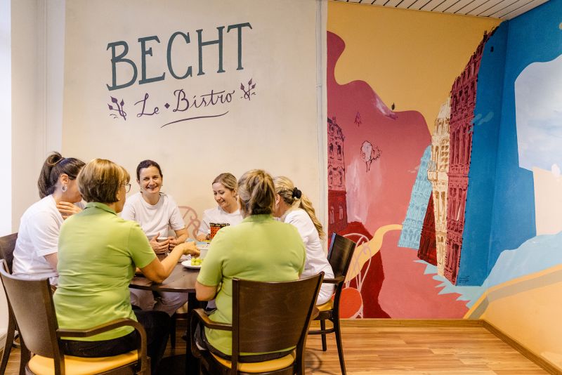 Eine Gruppe von Frauen sitzt an einem Tisch und unterhält sich in einem Bistro mit bunter Wandkunst und dem Schild "Becht Le Bistro".