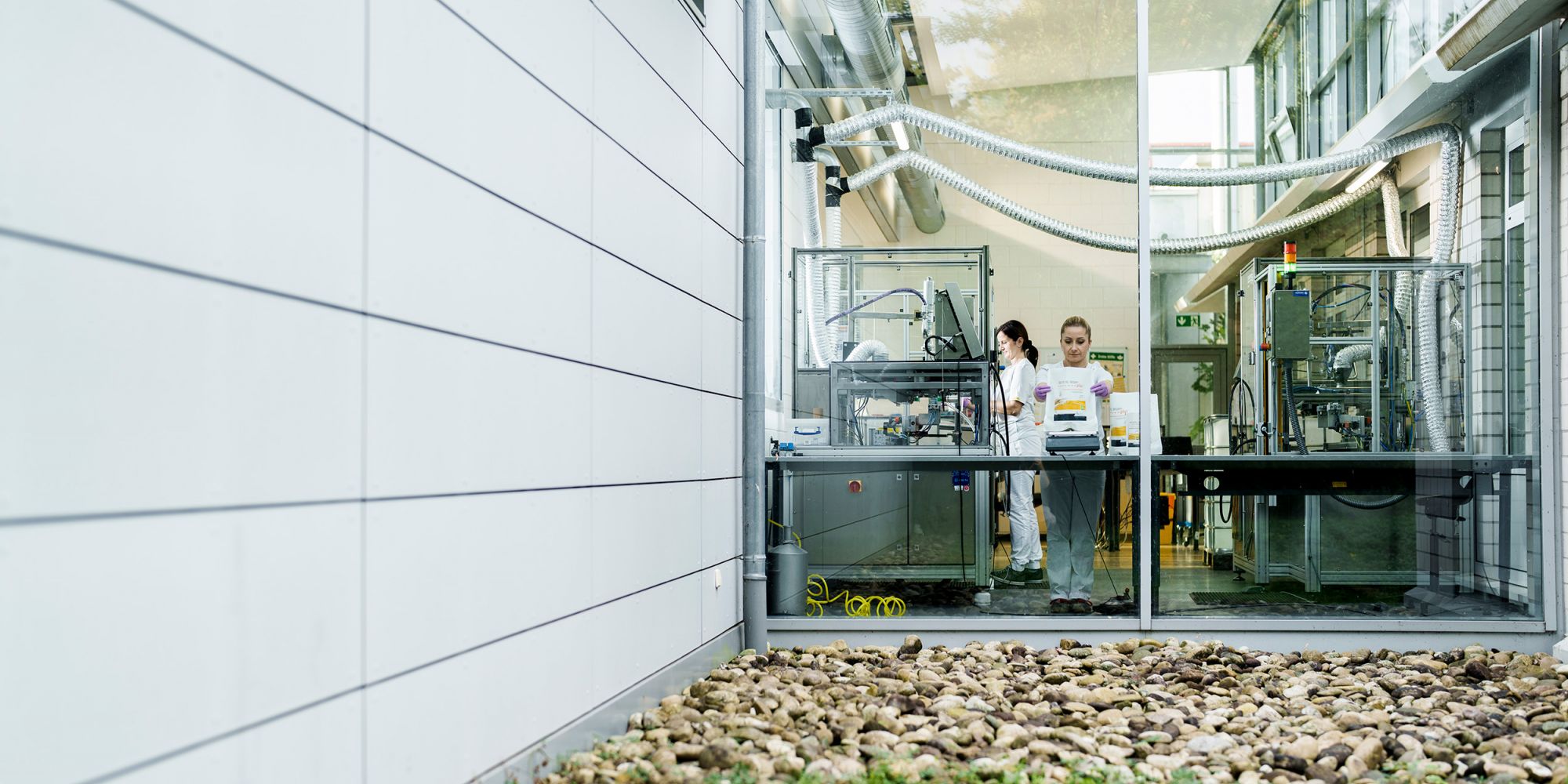 View through the glass façade into the modern production hall of Alfred Becht GmbH, where employees are working on machines.