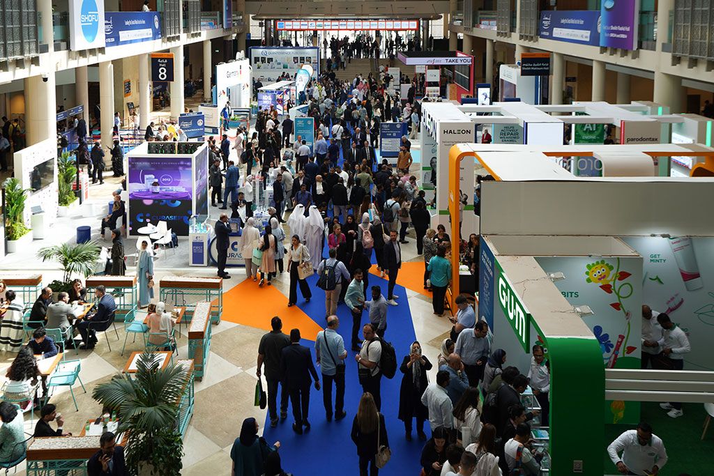 Eine belebte Messehalle mit zahlreichen Besuchern, die auf einem blau-orangefarbenen Bodenbelag zwischen verschiedenen Messeständen umhergehen.