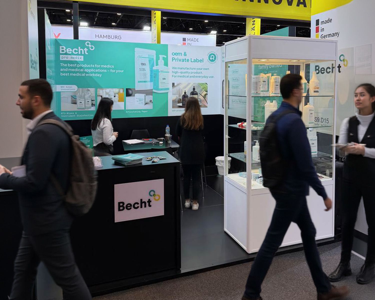 Ein belebter Messestand der Firma Becht mit schwarzem Tresen, einer großen Werbewand und einer Glasvitrine mit Produkten.