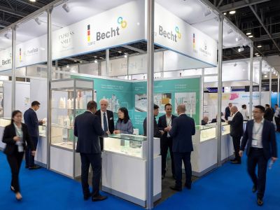 Ansicht eines hell beleuchteten Eck-Messestandes in Weiß und Türkis. Mehrere Gruppen von Geschäftsleuten in Anzügen führen Gespräche vor Glasvitrinen mit Produkten. Oben ist das Logo von Becht zu sehen.