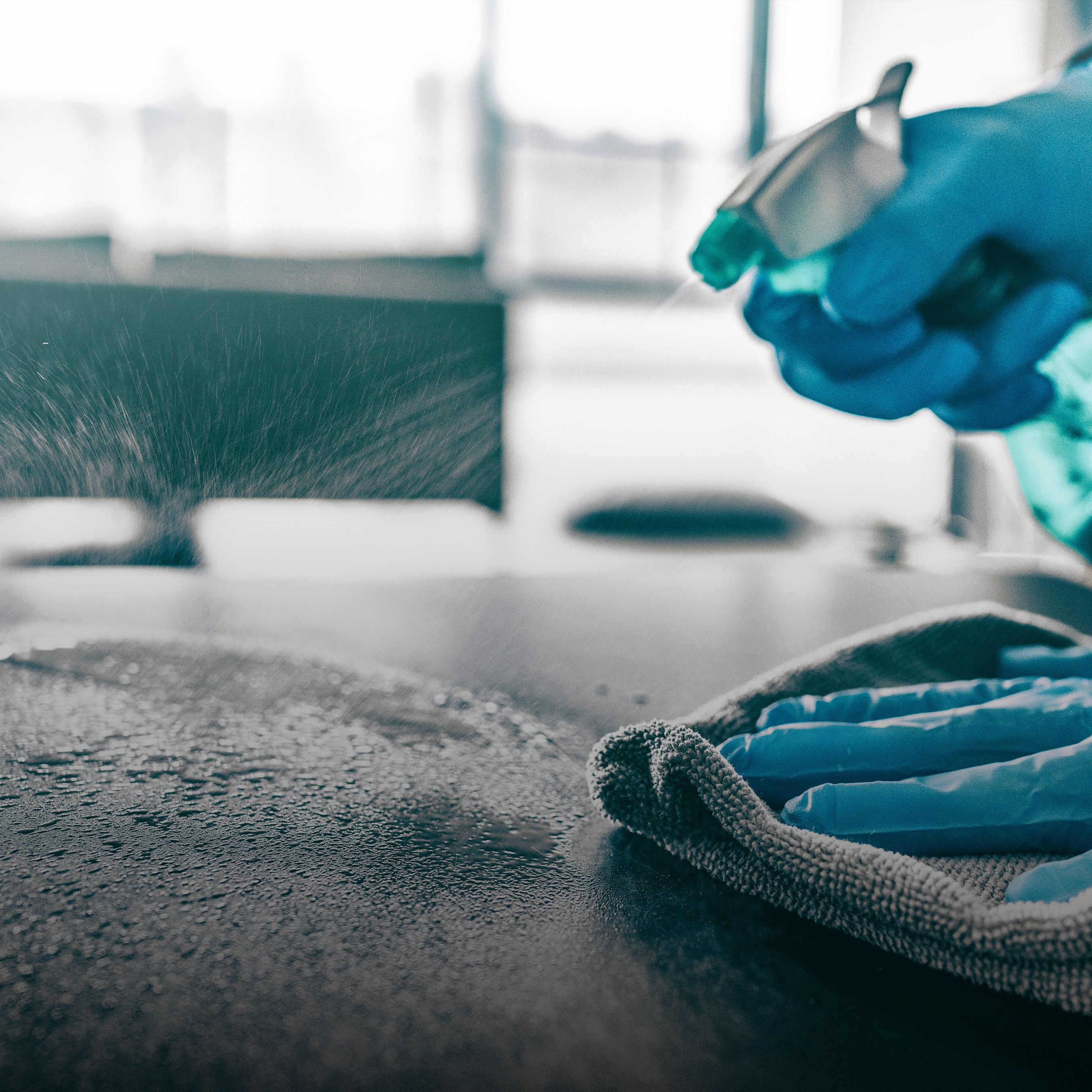 A gloved hand sprays disinfectant on a surface, wiping it clean with a cloth.