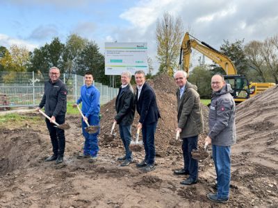 Sechs Männer stehen auf einer Baustelle und führen einen symbolischen Spatenstich durch. Im Hintergrund sind Erdhügel, ein Bagger und ein Bauschild zu sehen.