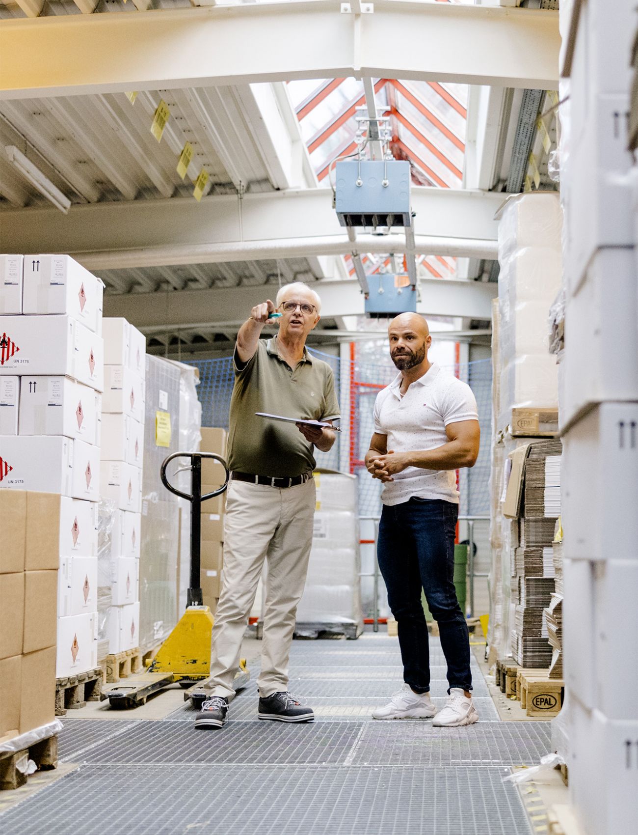 Two men in a warehouse full of stacked boxes, with one pointing while the other listens.