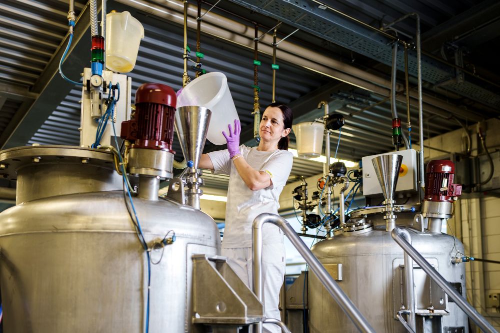 An employee of Alfred Becht GmbH works in production, pouring raw materials into a large stainless-steel tank.