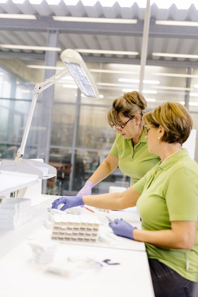 Zwei Frauen in grünen Polohemden und lila Handschuhen arbeiten sorgfältig in einem Labor. Eine Frau hantiert vorsichtig mit Materialien unter einer hellen Schreibtischlampe, während die andere assistiert. Auf dem Tisch sind ordentlich gestapelte Vorräte.