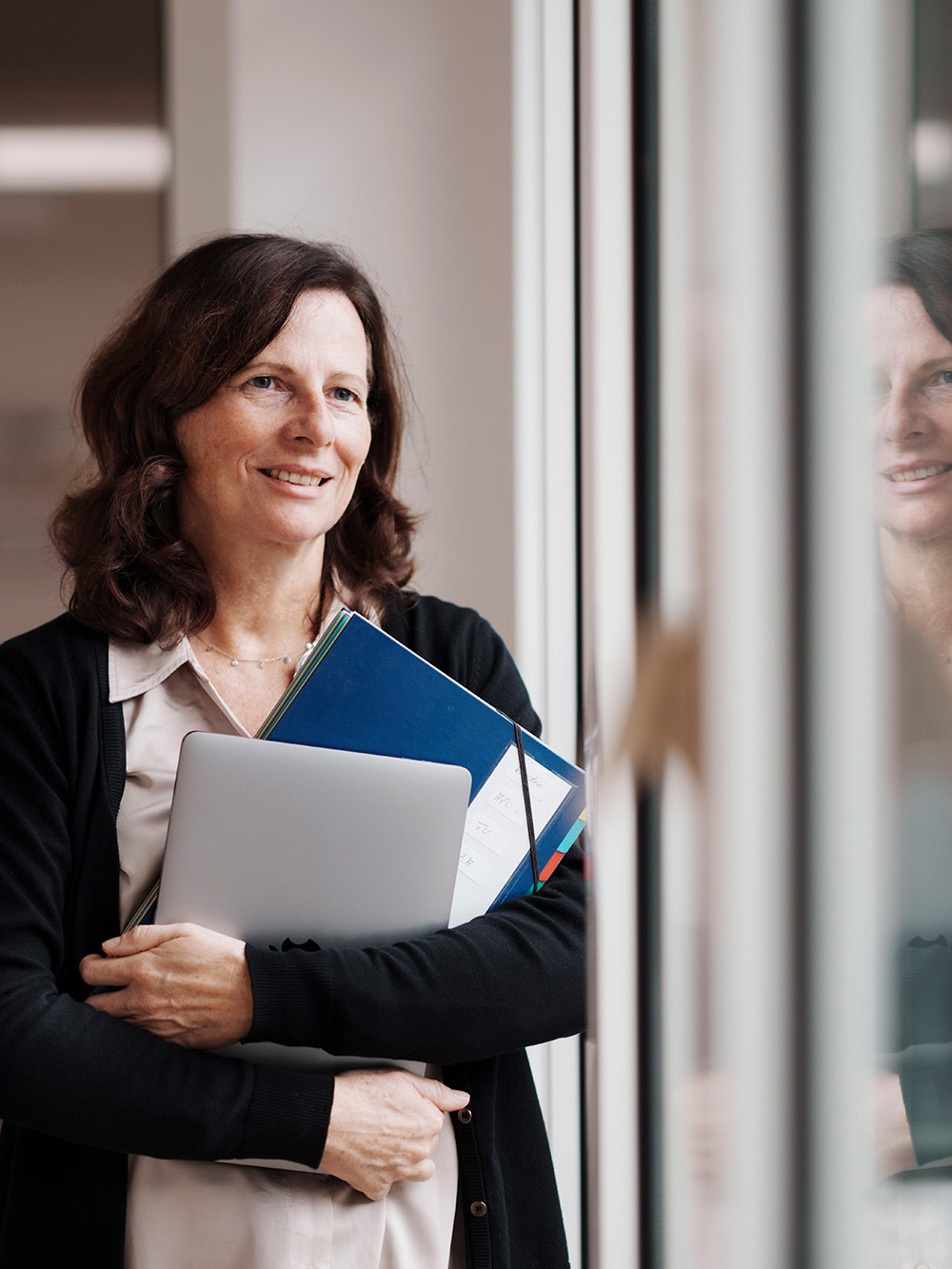 An employee of Alfred Becht GmbH stands by a window holding a laptop and documents, smiling.