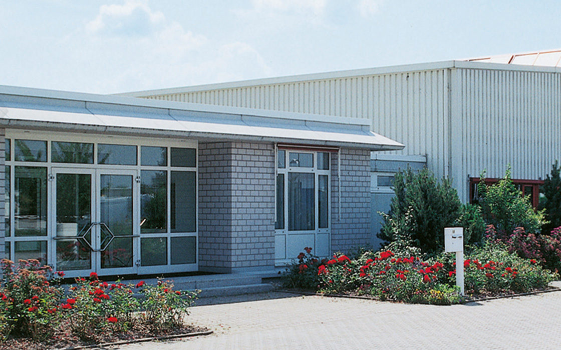 Alfred Becht company building with glass façade and adjacent production hall, surrounded by landscaped greenery and blooming roses.
