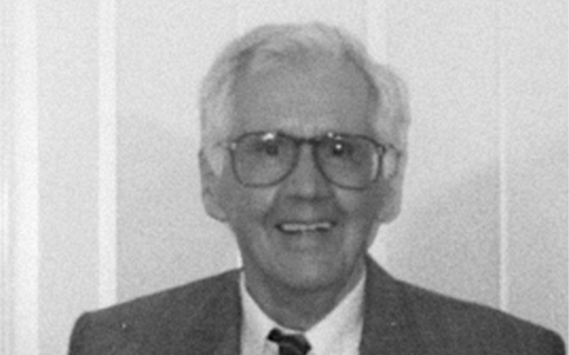 Black-and-white portrait of a man wearing glasses, a suit, and a tie, smiling warmly against a light background.