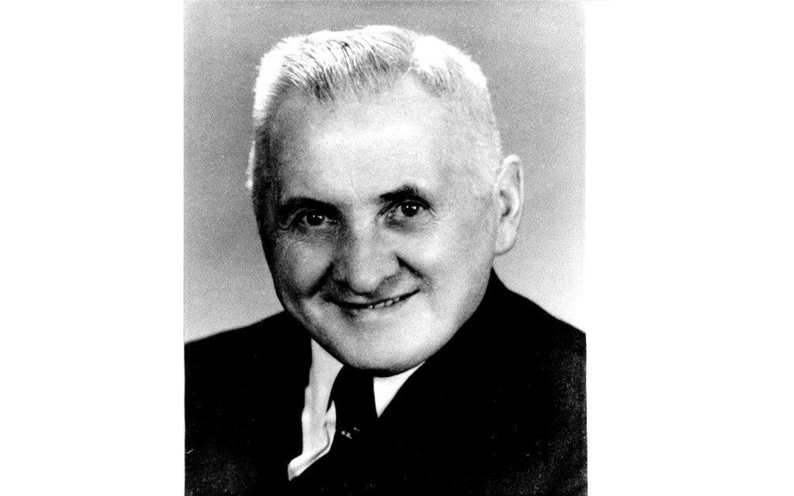 Black and white portrait of an elderly man with short light hair, wearing a suit and smiling warmly against a neutral background.